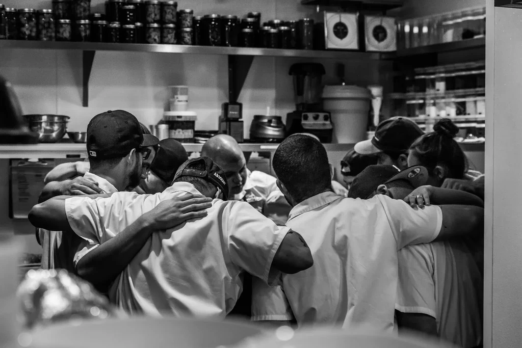 Professional kitchen staff huddle together, arms around shoulders.