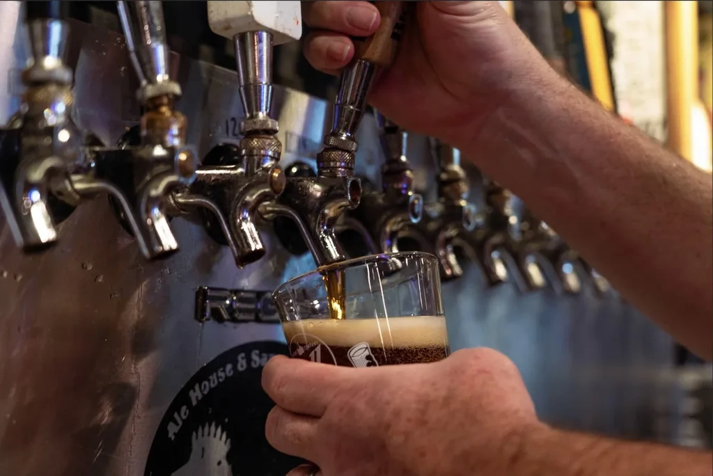 Hands pouring dark, frothy beer from a tap into a glass.