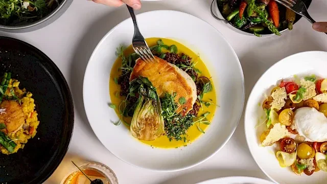 Hand forks pan-seared fish and gourmet dishes on a white table.