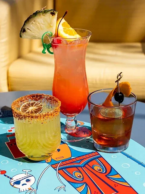 Three colorful cocktails on a blue, illustrative tiki-themed table.