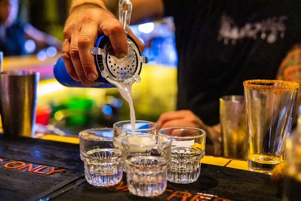 Bartender pours foamy liquid from blue shaker into shot glasses on bar counter.