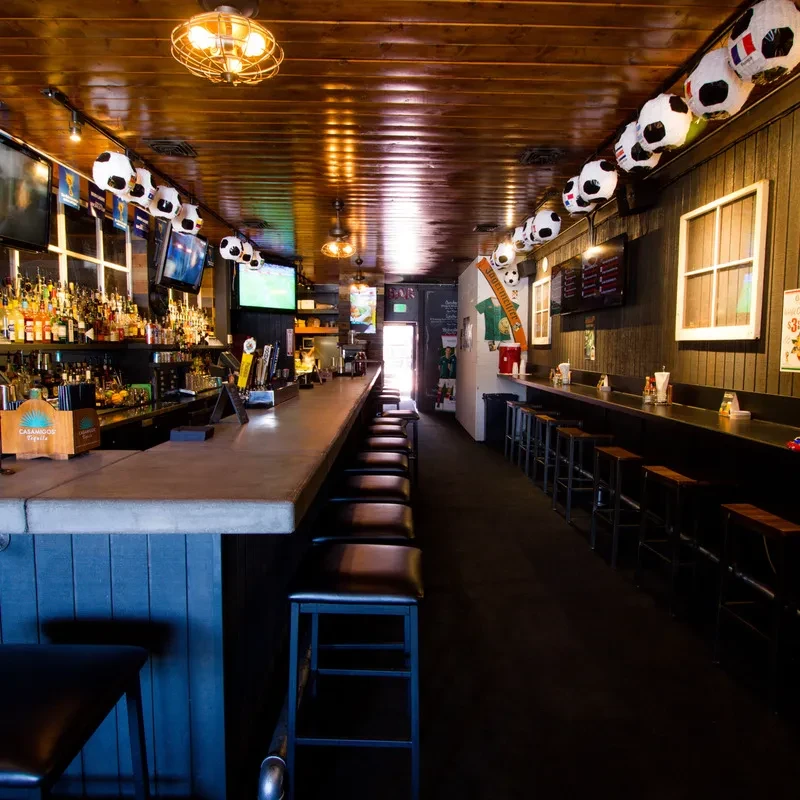 Long, empty bar with liquor bottles, multiple TVs, and soccer ball lights.
