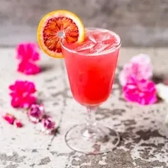 Red-pink cocktail with ice, blood orange slice, and pink flowers.