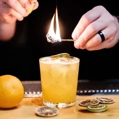 Hands ignite citrus zest, creating a bright flame over an orange cocktail.