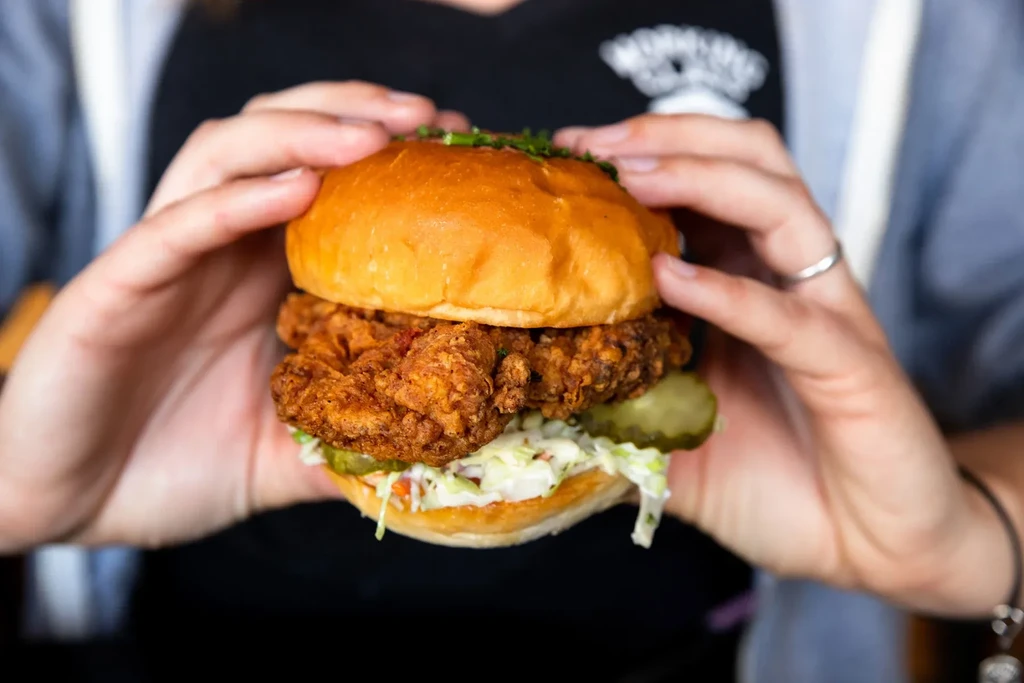 Hands holding a crispy fried chicken sandwich with coleslaw, dill pickles.