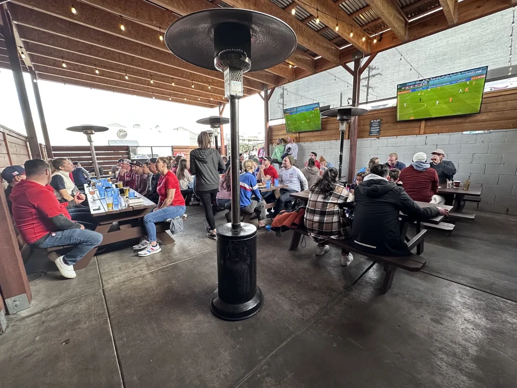 Semi-outdoor restaurant patio with patrons, picnic tables, sports on large TVs.