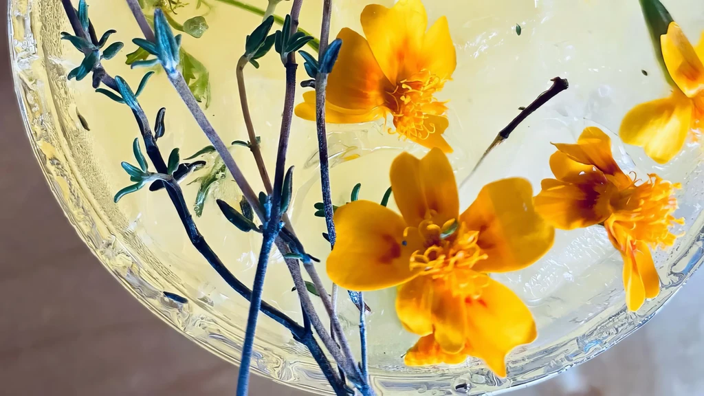 Clear cocktail with green herbs and yellow flowers over ice.