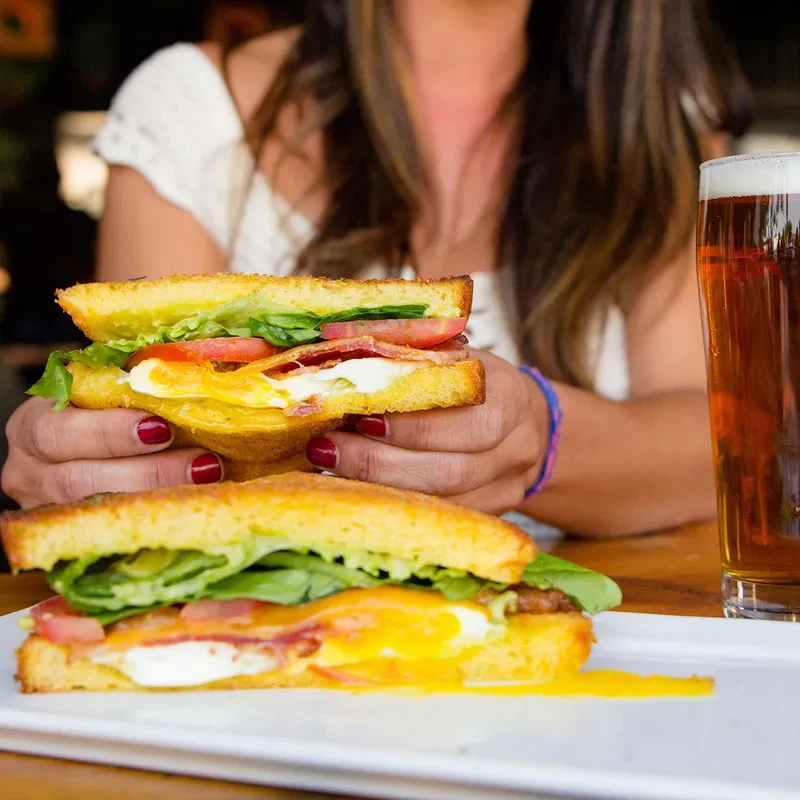 Hands holding a runny egg and bacon sandwich with beer.