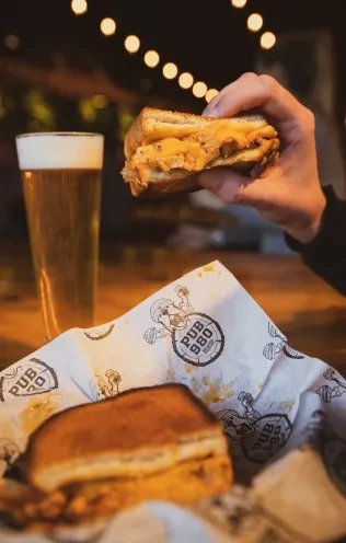 Hand holds cheesy grilled sandwich, beer, and half sandwich in pub.