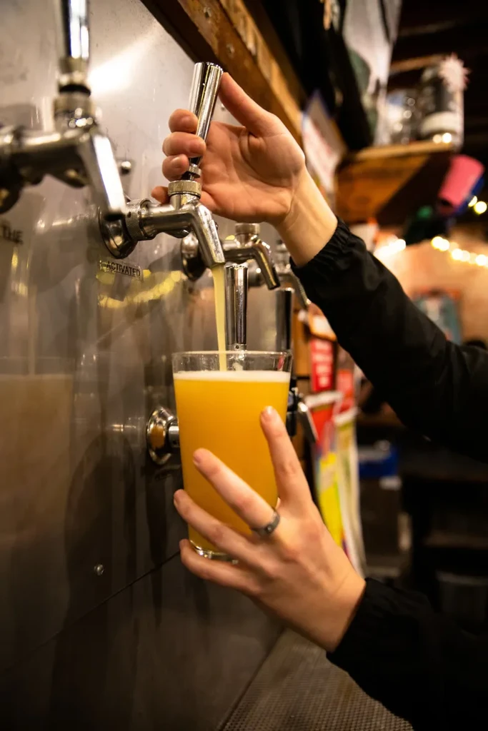 Hands pouring a golden drink from a chrome tap into a glass.