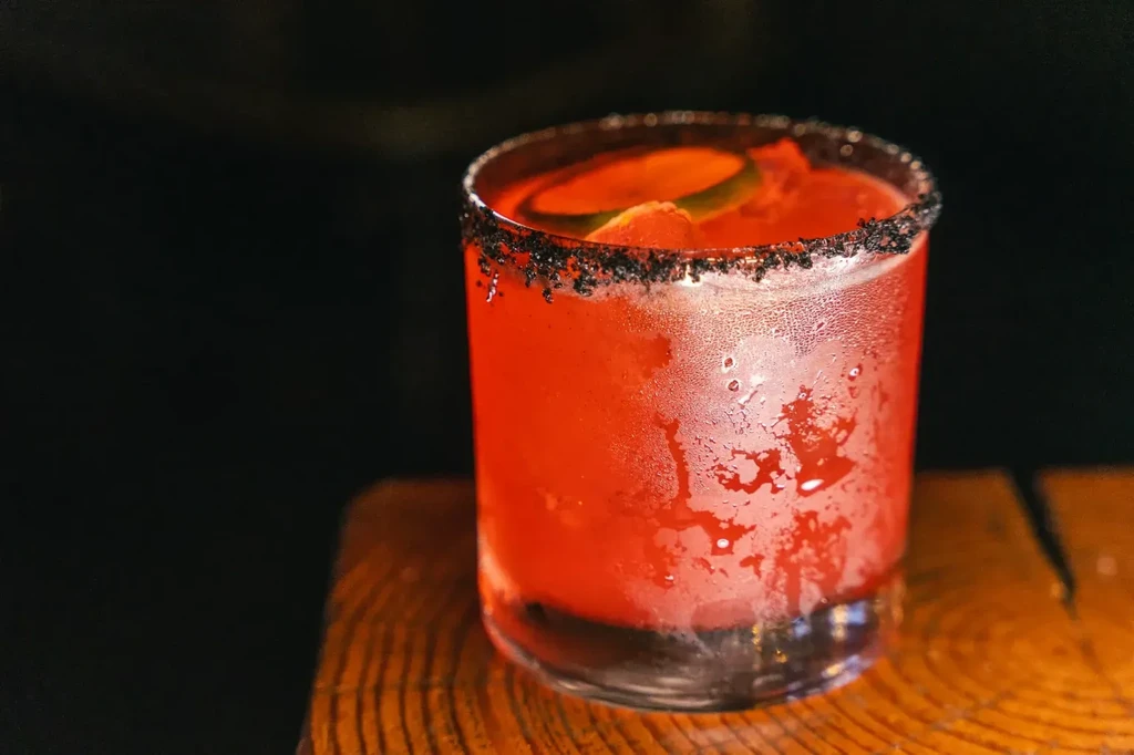 Frosted red cocktail with dark rim and citrus on wooden surface.