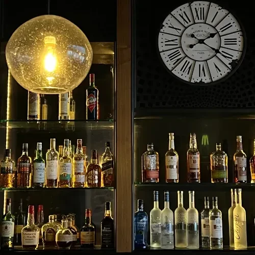 Well-lit bar with illuminated spirits, a pendant light, and a vintage wall clock.