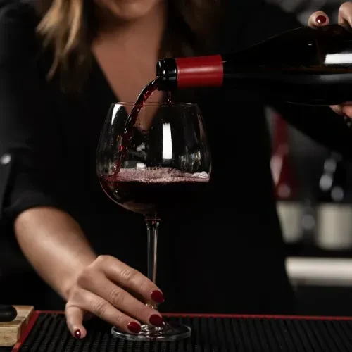 Hands pouring red wine from a dark bottle into a clear glass.