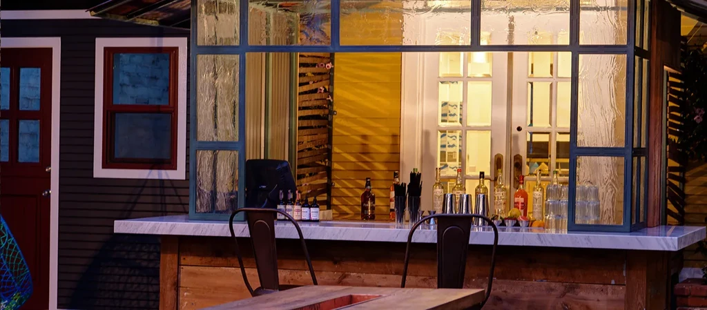 Outdoor bar with liquor, tools, two stools, and a red door.
