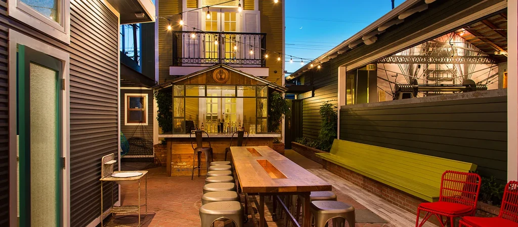 Inviting outdoor patio at dusk with communal table, bright bar, string lights.