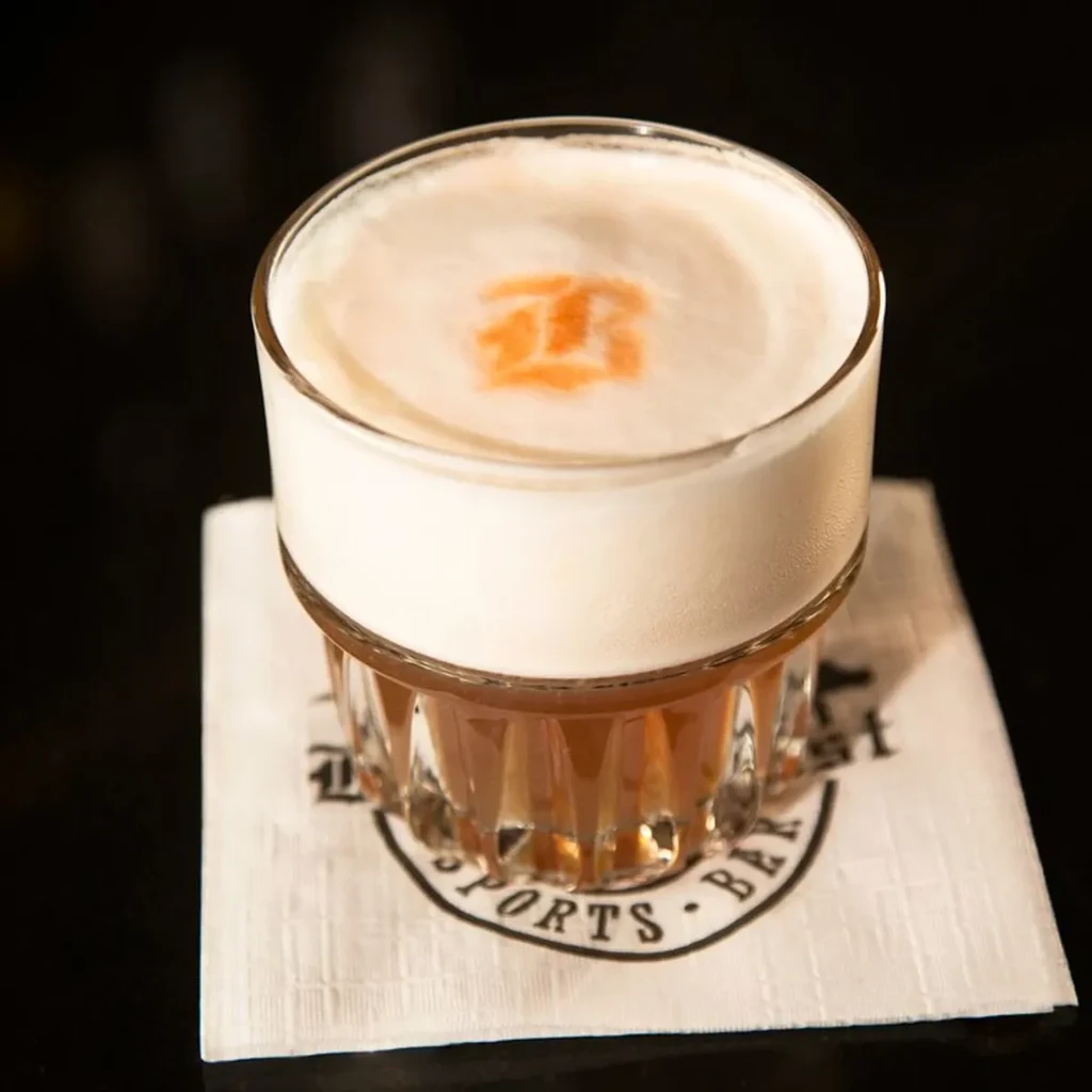 Amber cocktail with white foam and orange design in a ridged glass.