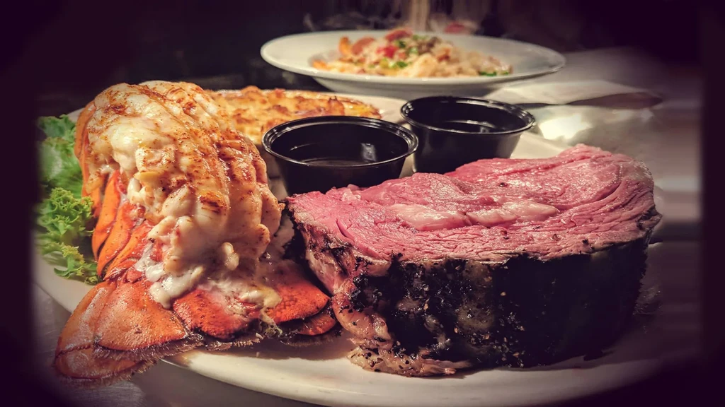 Seared prime rib, grilled lobster tail, and sauces on a white plate.