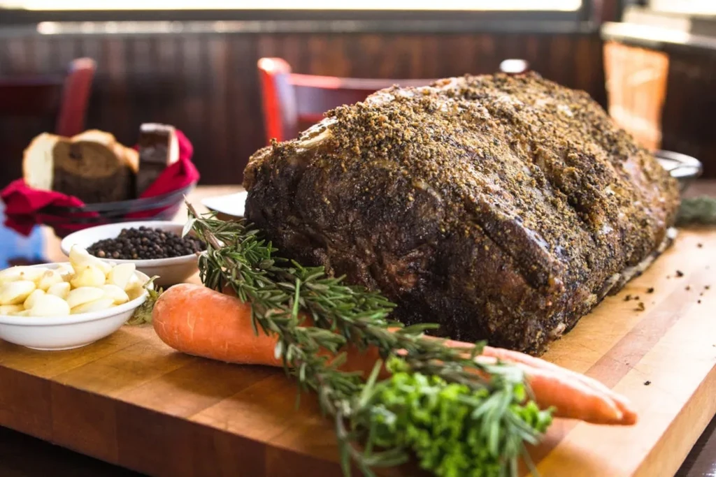 Large seasoned beef roast on a cutting board with herbs, carrots, garlic.