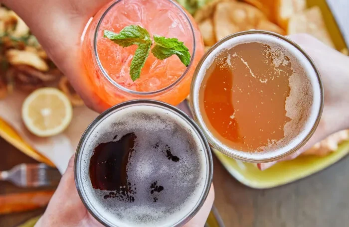 Three hands toast with a pink cocktail, amber beer, and dark beer.