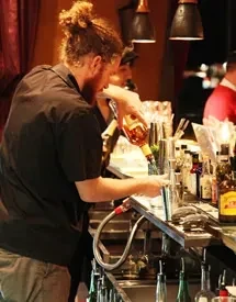 Bartender with bunned hair pours a drink in a dimly lit bar.