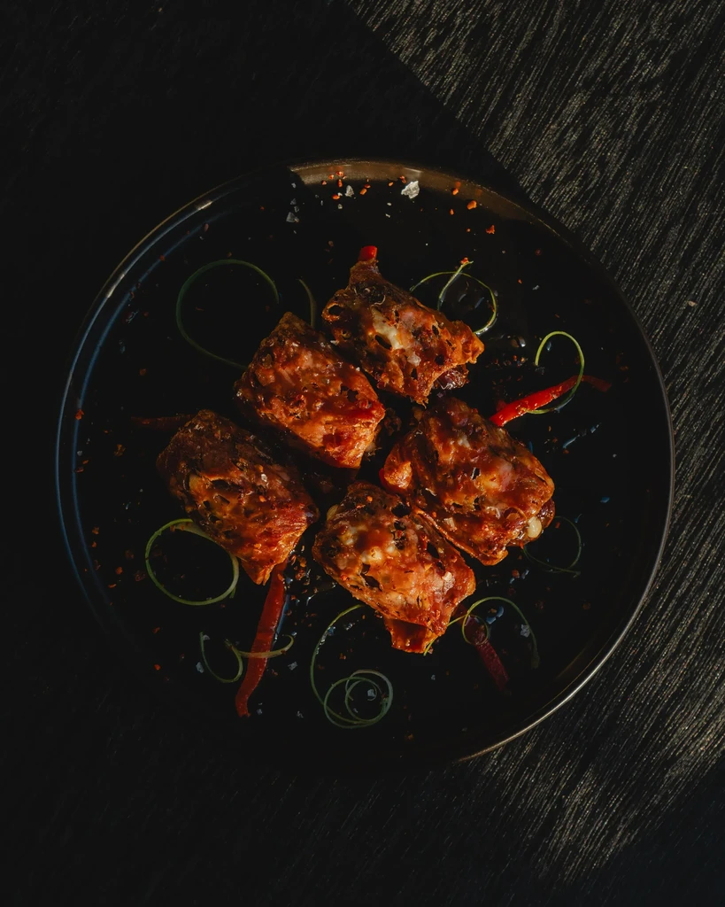 Dark plate with five pieces of glazed, seasoned meat, herbs, and chilies.