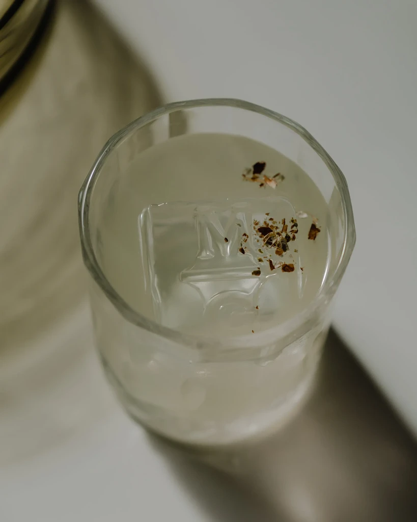 Light cocktail in faceted glass, 'M' embossed ice, dark granular garnish.