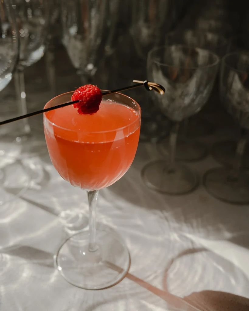 Vibrant coral-red cocktail in stemmed glass with raspberry garnish.