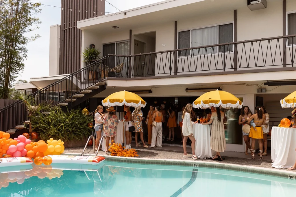 Vibrant outdoor poolside gathering with people, tables, umbrellas, and balloons.