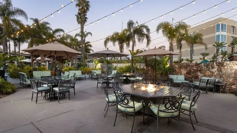Tropical outdoor patio at dusk with fire pit, palm trees, and string lights.