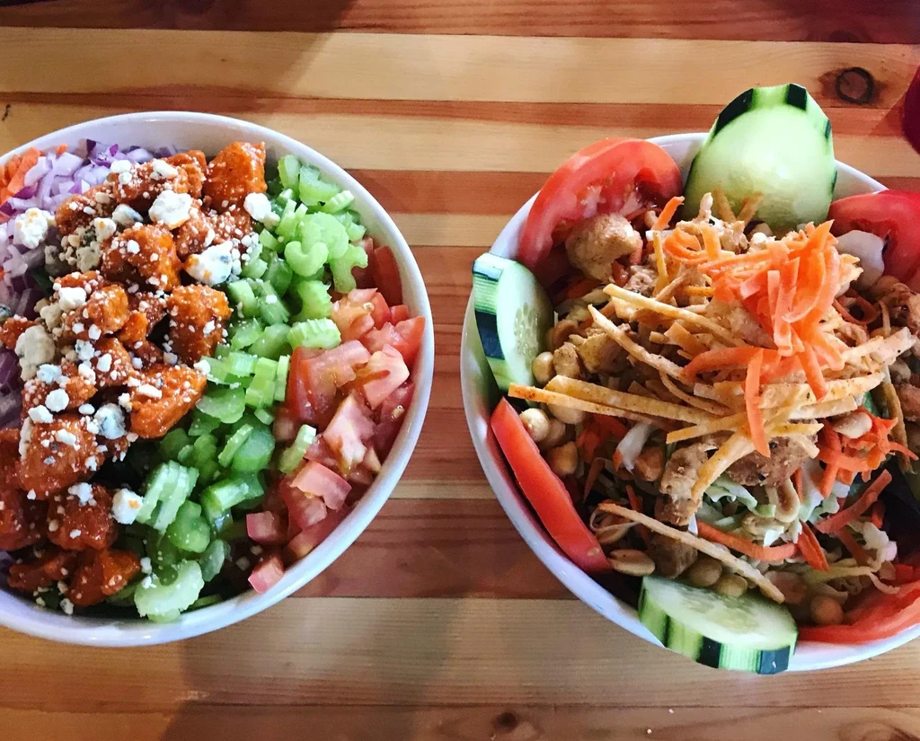 Two colorful salads, buffalo chicken and Asian-inspired, on a wooden table.