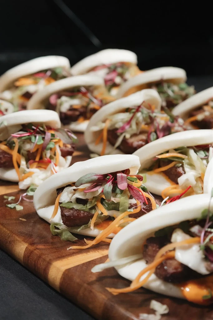 Open bao buns filled with meat, carrots, lettuce, microgreens, and sauces.