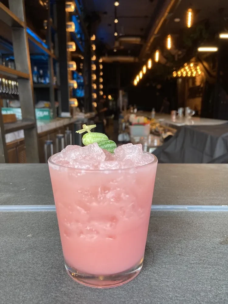 Pink cocktail with crushed ice and cucamelon garnish at a bar.