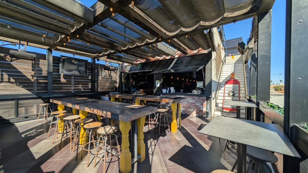 Sunny outdoor bar area with wooden tables, yellow bases, Skrewball mural.