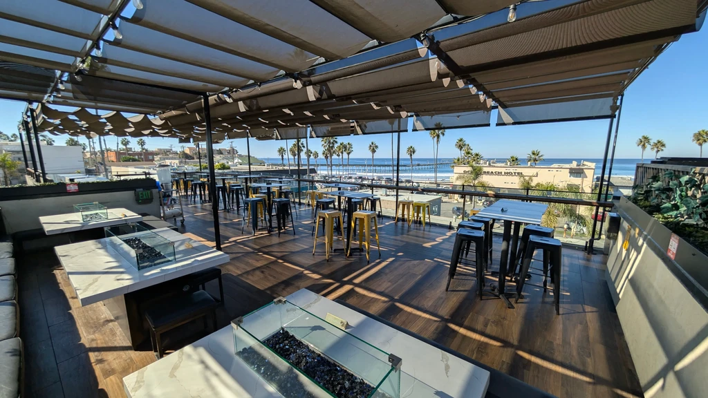 Sunny rooftop patio with ocean view, lounge seating, and high-top tables.