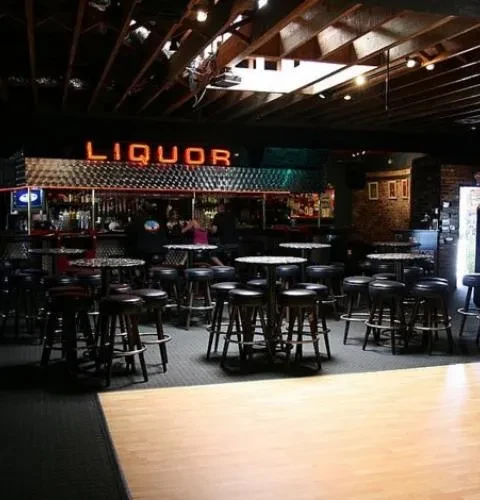 Dimly lit bar interior with "LIQUOR" neon sign and high-top tables.