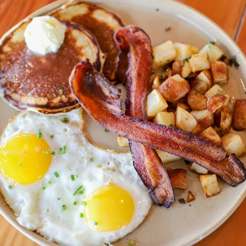 Breakfast plate with pancakes, crispy bacon, roasted potatoes, and sunny-side-up eggs.