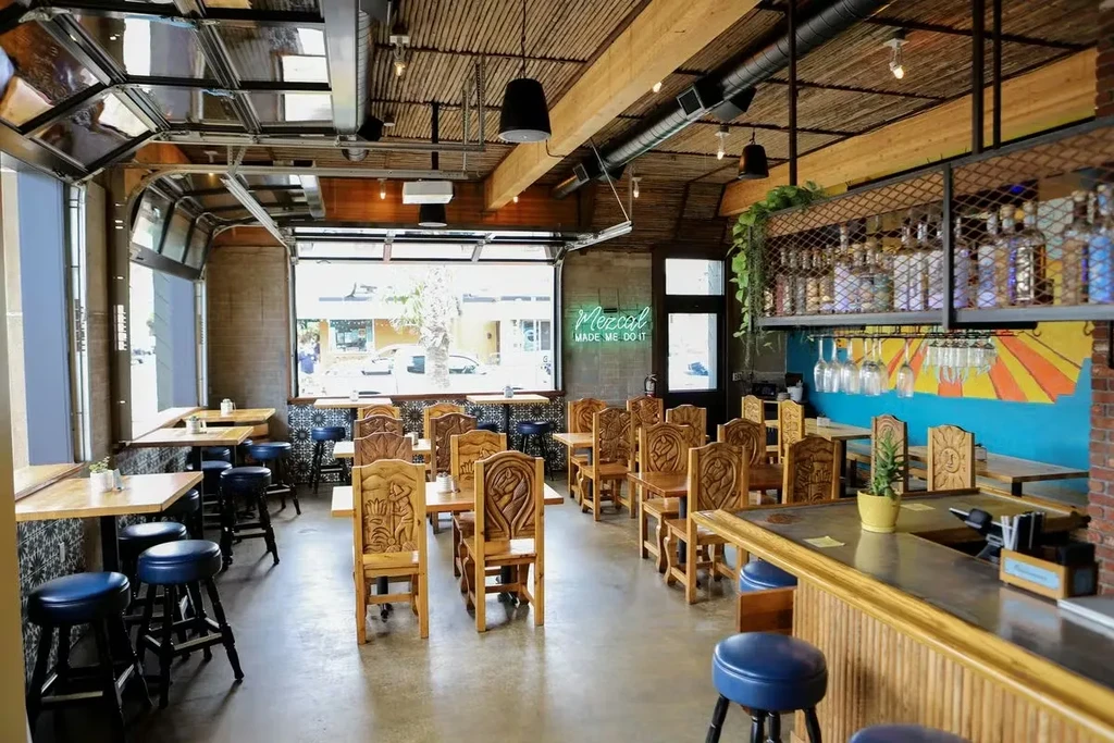 Restaurant interior with dining tables, bar, and a neon "Mezcal" sign.