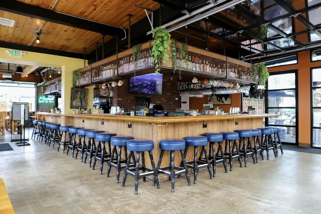 Bright, modern bar with L-shaped wood counter, blue stools, and liquor.