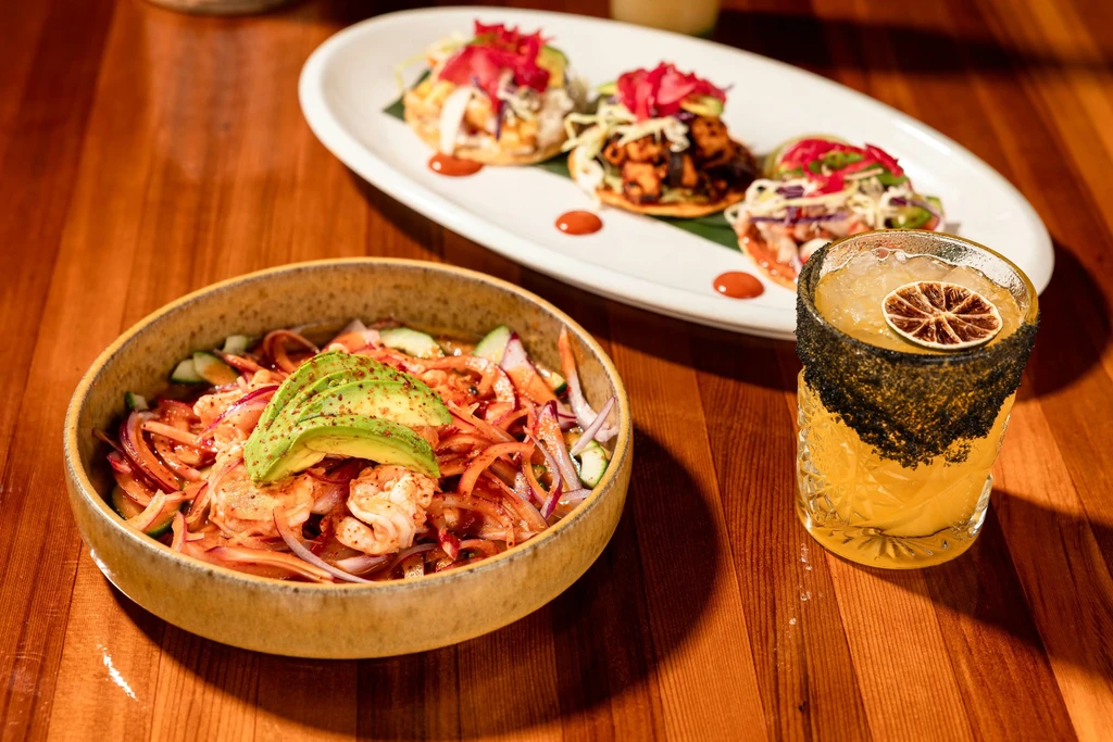 Ceviche bowl, cocktail, and mini tostadas on a wooden table.