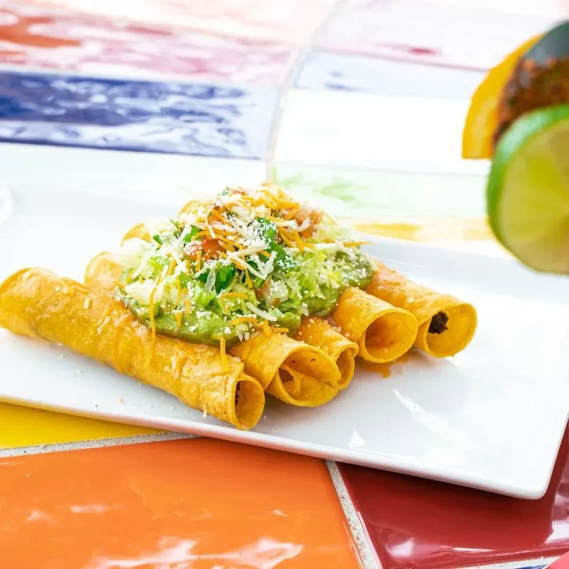 Five taquitos topped with guacamole, lettuce, tomato, and cheese on a plate.