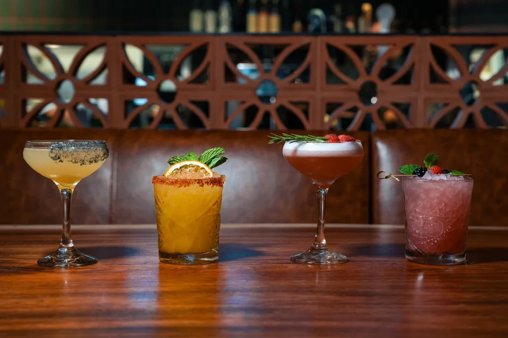 Four elaborately garnished cocktails on a wooden bar.