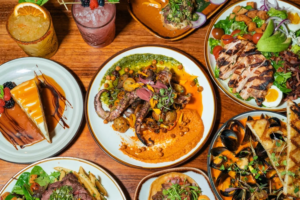 Diverse spread of grilled octopus, steak, salad, mussels, cheesecake, and cocktails.