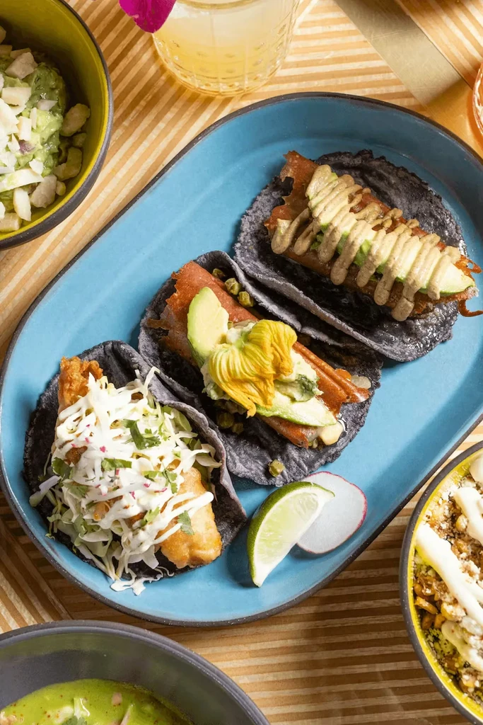 Three distinct tacos on an oval platter surrounded by vibrant side dishes.
