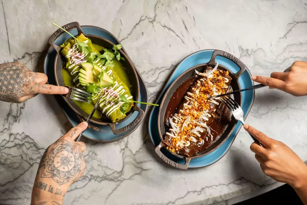 Overhead: Two diners' hands eating rolled dishes with green and mole sauces.