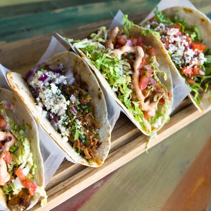Five colorful tacos with various fillings diagonally arranged on a wooden board.