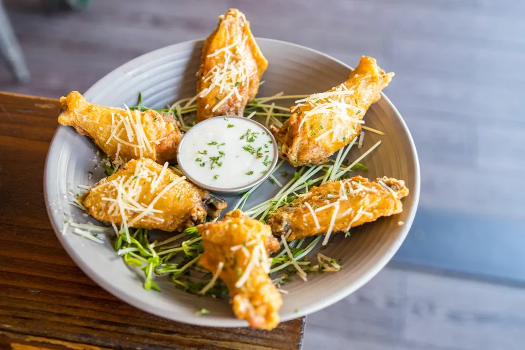 Plate of six Parmesan chicken wings with dipping sauce and green sprouts.