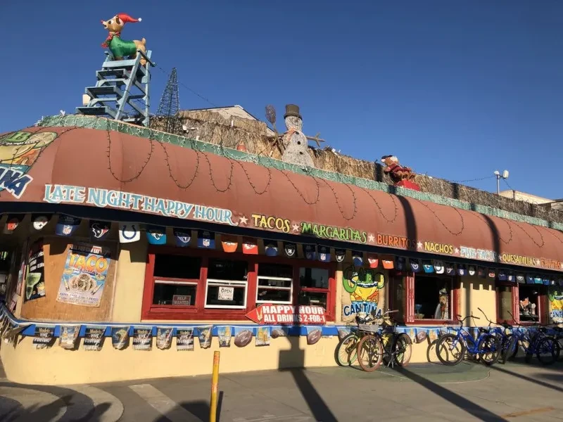 Kitschy restaurant exterior with festive roof decor, cactus, and food signs.