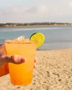 Hand holding orange frozen drink, lime, seasoned rim, beach background.