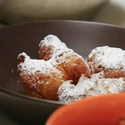 Irregular golden-brown fried dough with powdered sugar in a dark bowl.