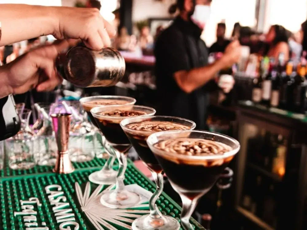 Hands pour frothy cocktails into four martini glasses on a Casamigos mat.